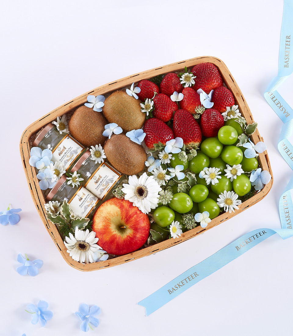 Rectangular woven fruit basket with fresh kiwis, strawberries, green plums, red apple, wellness bottles and white daisies