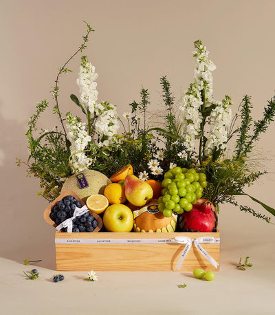 Premium wooden fruit gift box with fresh cantaloupe, grapes, berries, and citrus, surrounded by white stock flowers and lush