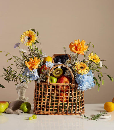Wicker basket filled with fresh fruits and vibrant yellow gerberas, orange roses, and blue hydrangeas for gifting