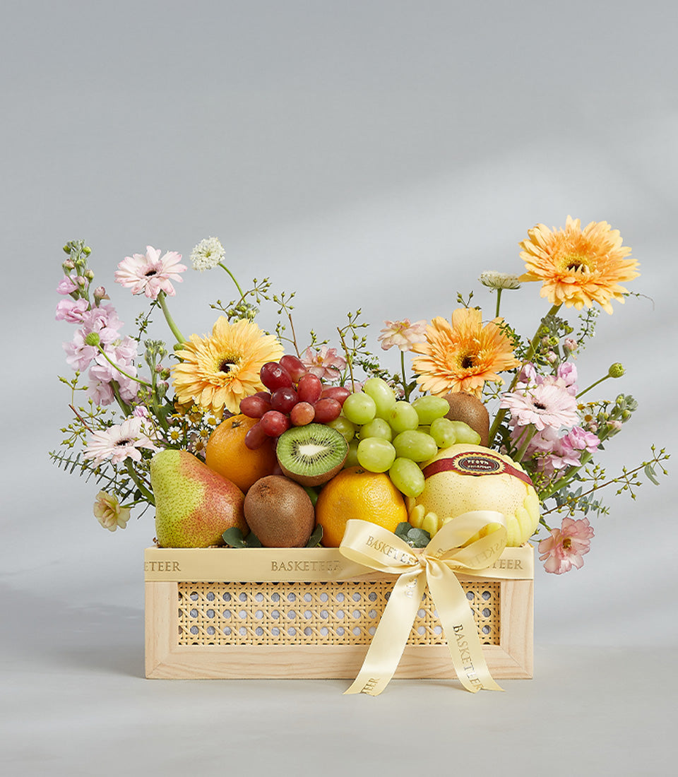 Light wooden crate with fresh red and green grapes, kiwis, oranges, pears, melon, and yellow-orange-pink gerbera daisies