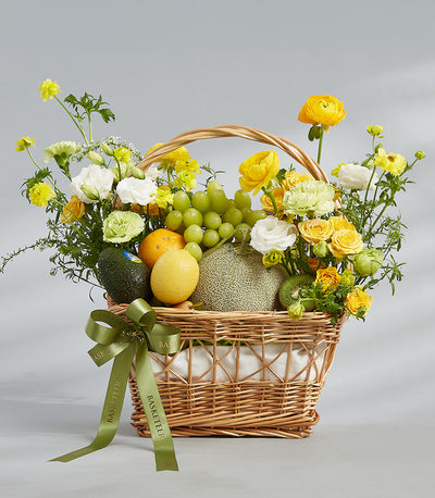 Wicker basket with fresh tropical fruits, yellow roses, and cream ranunculus with green satin bow