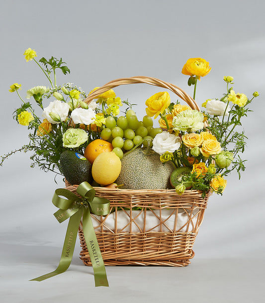 Wicker basket with fresh tropical fruits, yellow roses, and cream ranunculus with green satin bow