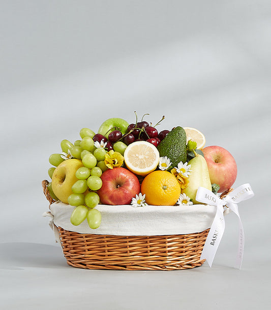 Woven wicker fruit basket with fresh grapes, apples, orange, cherries, and avocado, decorated with white flowers and ribbon