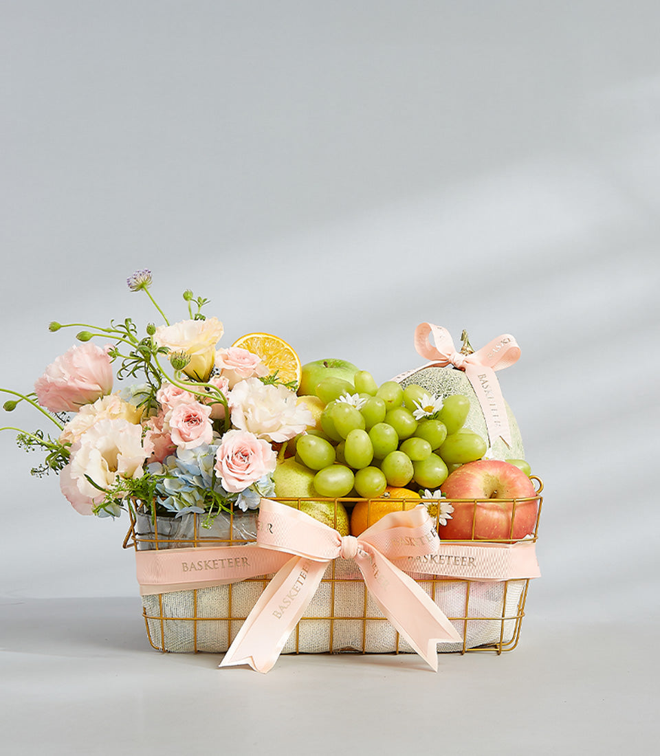 Pastel Harmony fruit basket with pink roses, hydrangeas, and fresh tropical fruits in gold wire mesh with pink ribbon bow