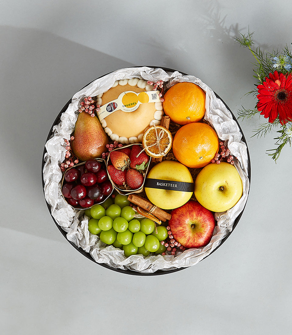 Deluxe fresh fruit gift basket with artisan dessert, cherries, strawberries, apples, pears, and grapes on white crinkle paper