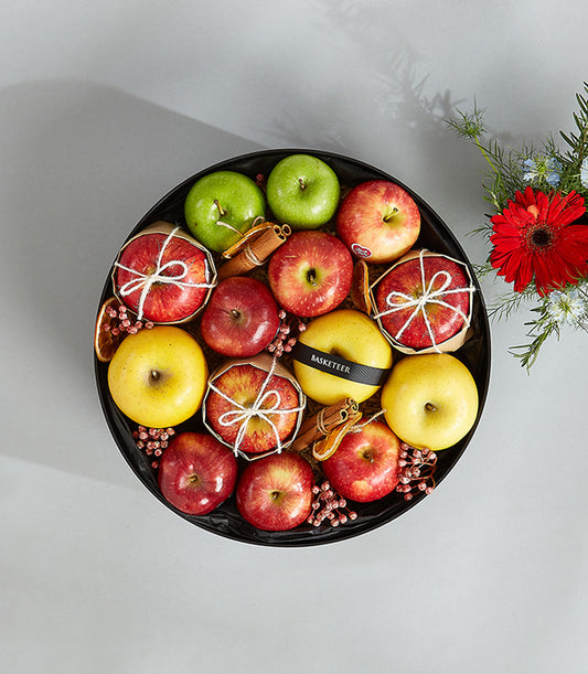 Elegant round black gift tray with green, red, and yellow apples, cinnamon sticks, dried citrus, and fresh red gerbera daisy