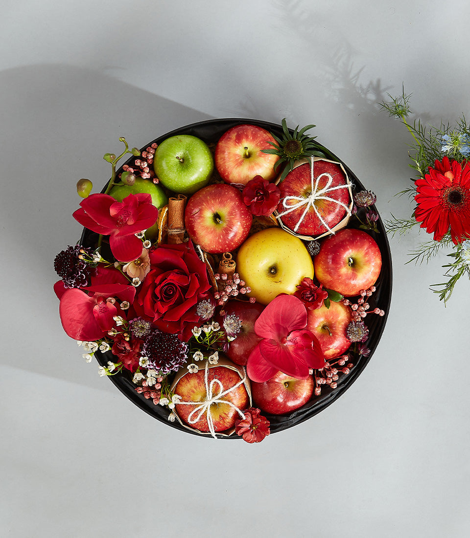 Romantic red fruit basket with red roses, orchids, apples, and cinnamon sticks in round black tray