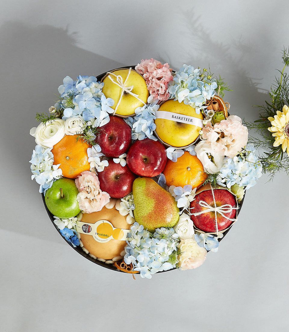 Elegant fruit and hydrangea gift basket with apples, oranges, and blue hydrangeas in round black container