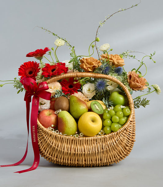 Woven wicker basket with fresh seasonal fruits, red apples, pears, kiwis, grapes, and vibrant red gerbera daisies, peach