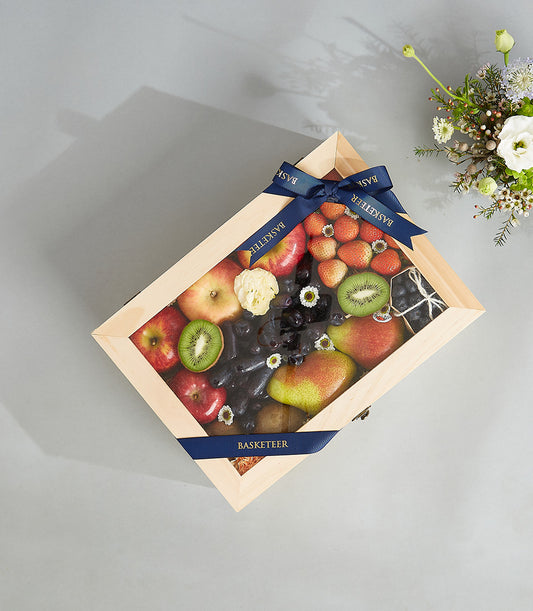 Light wooden diamond-shaped fruit box with clear lid holding fresh apples, kiwis, grapes, strawberries and pears, tied with