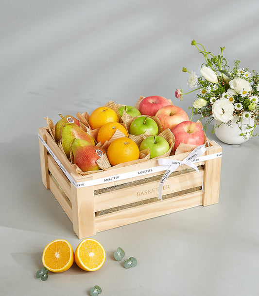 Rustic wooden crate filled with fresh red apples, green apples, oranges, and pears, wrapped individually in kraft paper