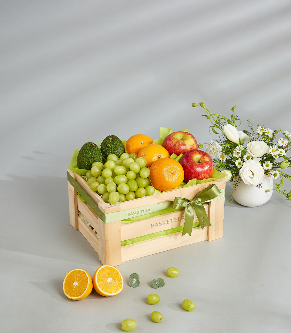 Wooden crate filled with fresh grapes, avocados, oranges, and apples, tied with green ribbon bow