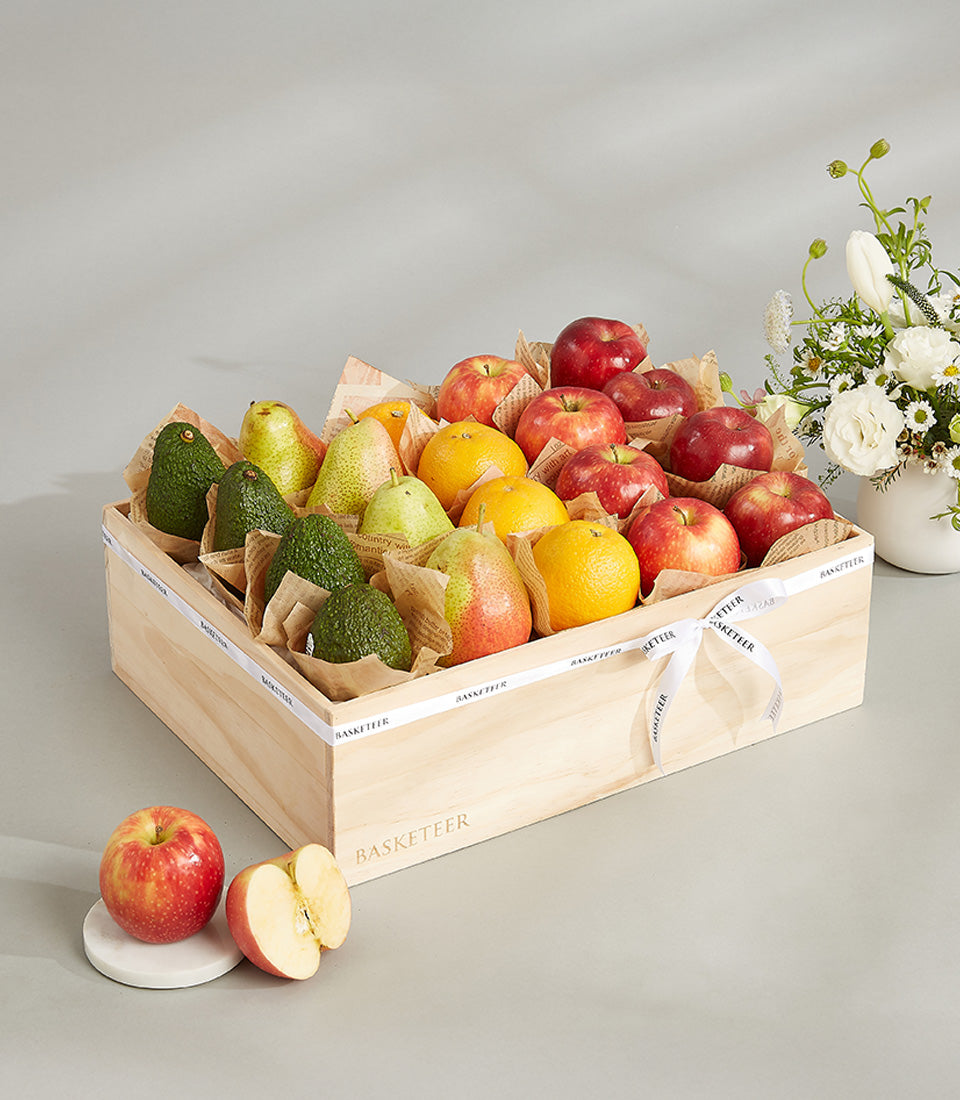 Light wooden crate filled with fresh apples, pears, oranges, and avocados, tied with white Basketeer ribbon