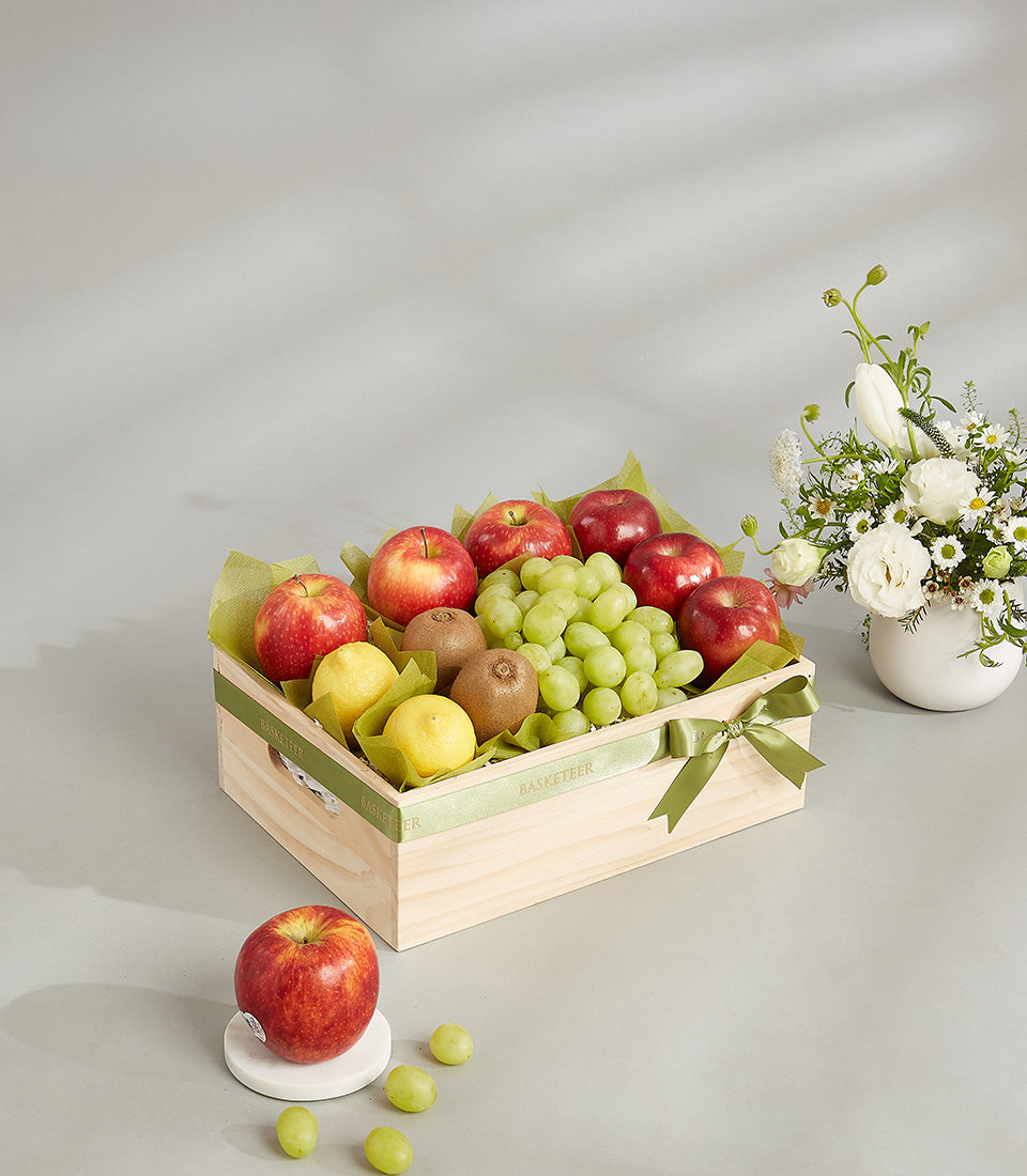 Wooden fruit gift crate with red apples, green grapes, lemons and kiwis on tissue paper with green ribbon bow