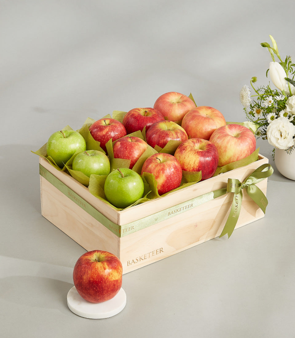 Wooden crate of premium red and green apples with white flowers in ceramic vase, luxury fruit gift hamper