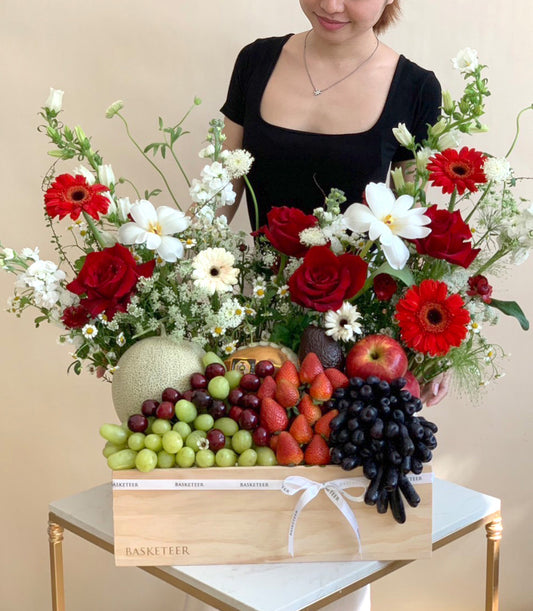 Wooden gift box filled with fresh tropical fruits and vibrant red roses, gerberas with white daisies