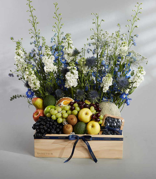 Luxury wooden fruit box with fresh grapes, apples, pears, and kiwis topped with white and blue delphinium flowers