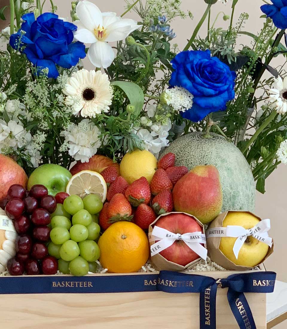Luxury blue roses and fresh fruit gift basket with wrapped apples, berries, and citrus in wooden crate