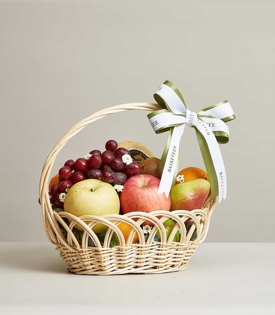 Light brown woven rattan basket with fresh red grapes, pears, apples, oranges and kiwis, white flower accents and