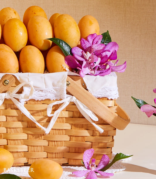 Woven picnic basket filled with golden mangoes, purple speckled orchids, and white lace trim with ribbon bows