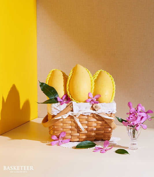 Premium Thai mango fruit basket with yellow protective netting, woven wicker, white ribbon, and fresh purple orchids