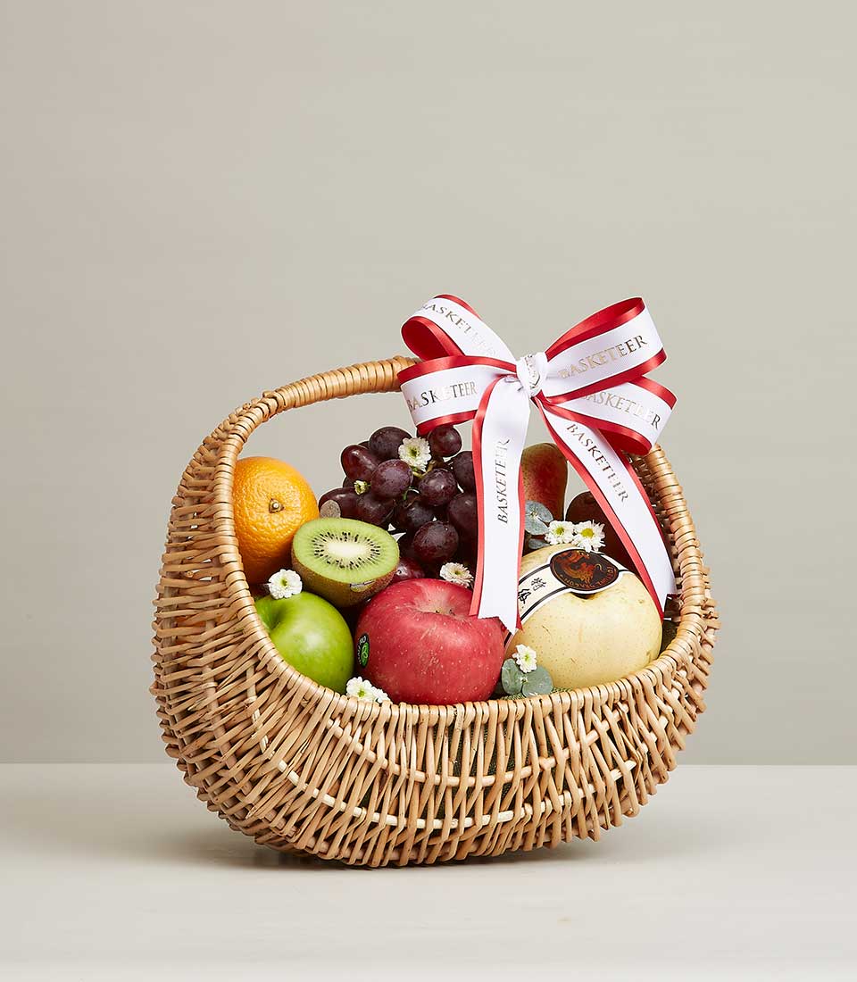 Wicker fruit basket with fresh oranges, apples, grapes, kiwi, and pear, tied with festive ribbon