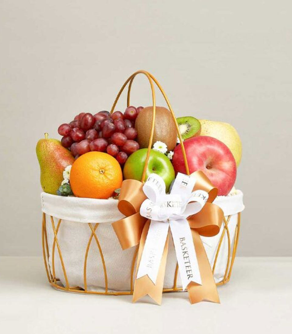 Gold-framed fruit basket with fresh grapes, kiwis, apples, oranges, pears, and white flowers, white and gold ribbon bow
