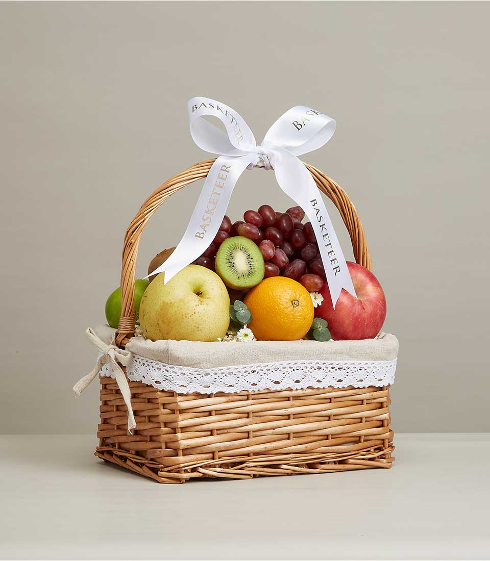 Woven wicker fruit basket filled with fresh grapes, pear, apple, orange and kiwi, lined with beige fabric and white lace trim
