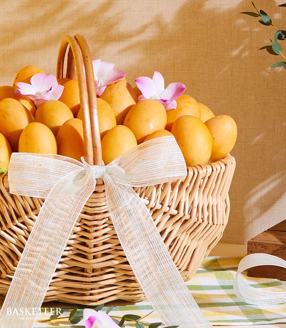 Wicker basket of ripe golden mangoes with pink and white orchids and light blue striped ribbon bow