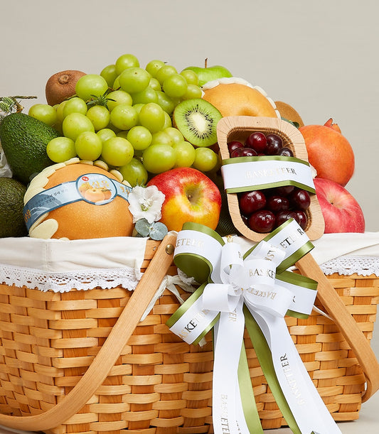 Luxury fruit basket with green grapes, avocados, kiwi, and pomegranate in woven wood with white lace and white-olive ribbon