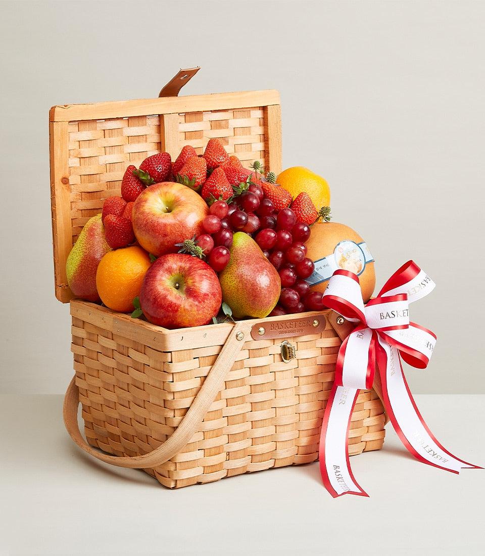 Woven wooden picnic basket filled with fresh red apples, pears, oranges, grapes, and strawberries, tied with white and red
