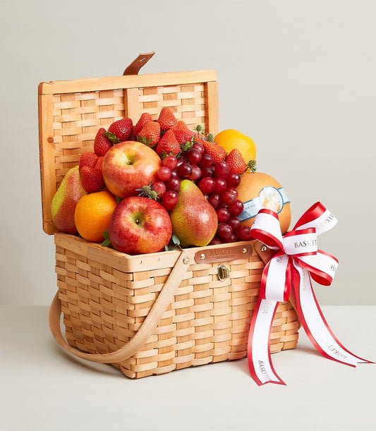 Woven wooden picnic basket filled with fresh red apples, pears, oranges, grapes, and strawberries, tied with white and red