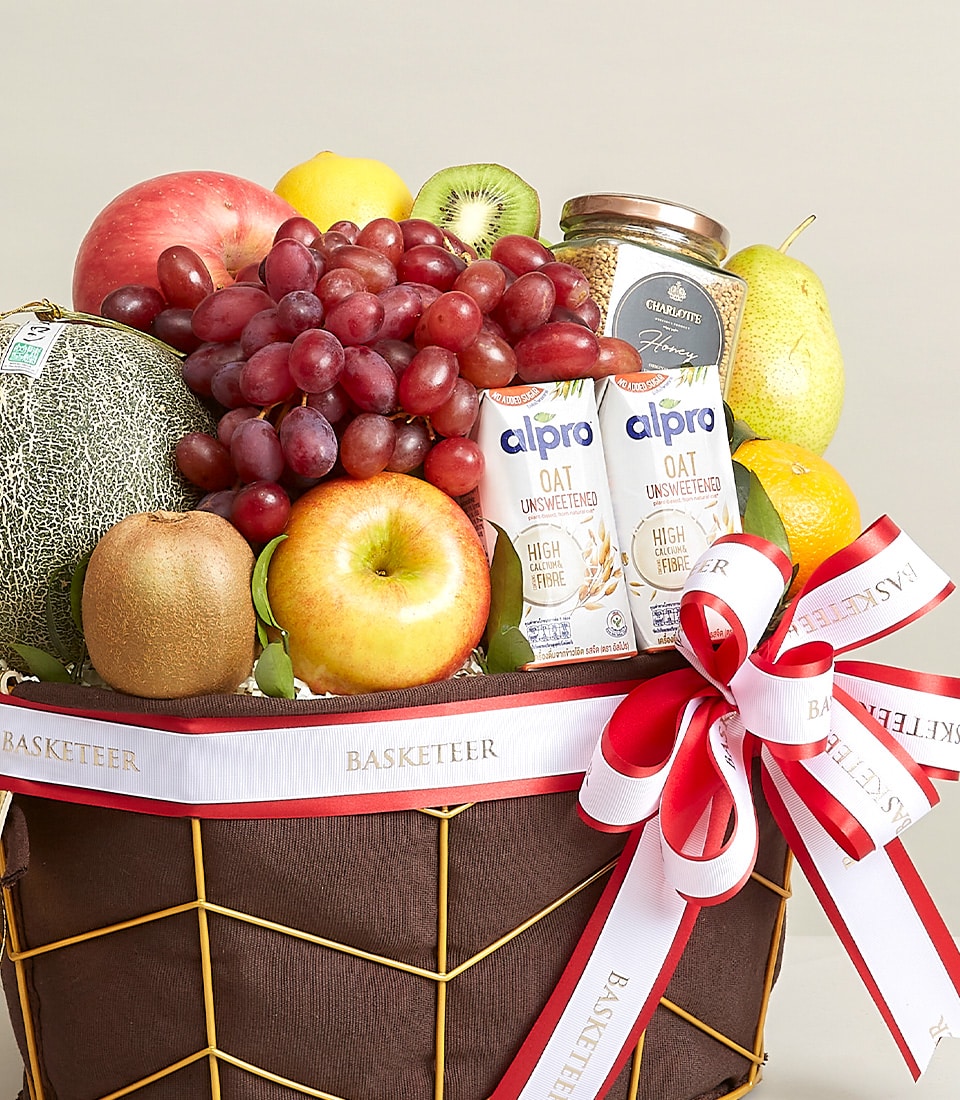 Dark brown fabric luxury fruit basket with gold wire frame, red and white ribbon bow, fresh tropical fruits, Alpro oat milk,