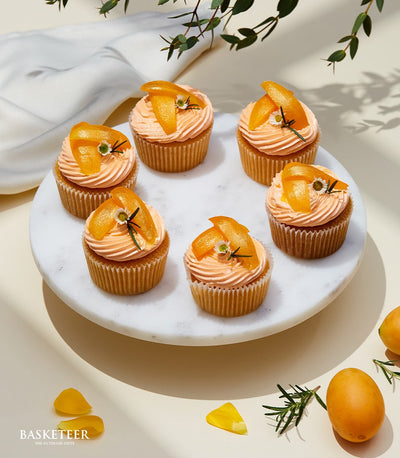 Six orange-frosted cupcakes with candied citrus, edible flowers, and fresh herbs on marble serving tray