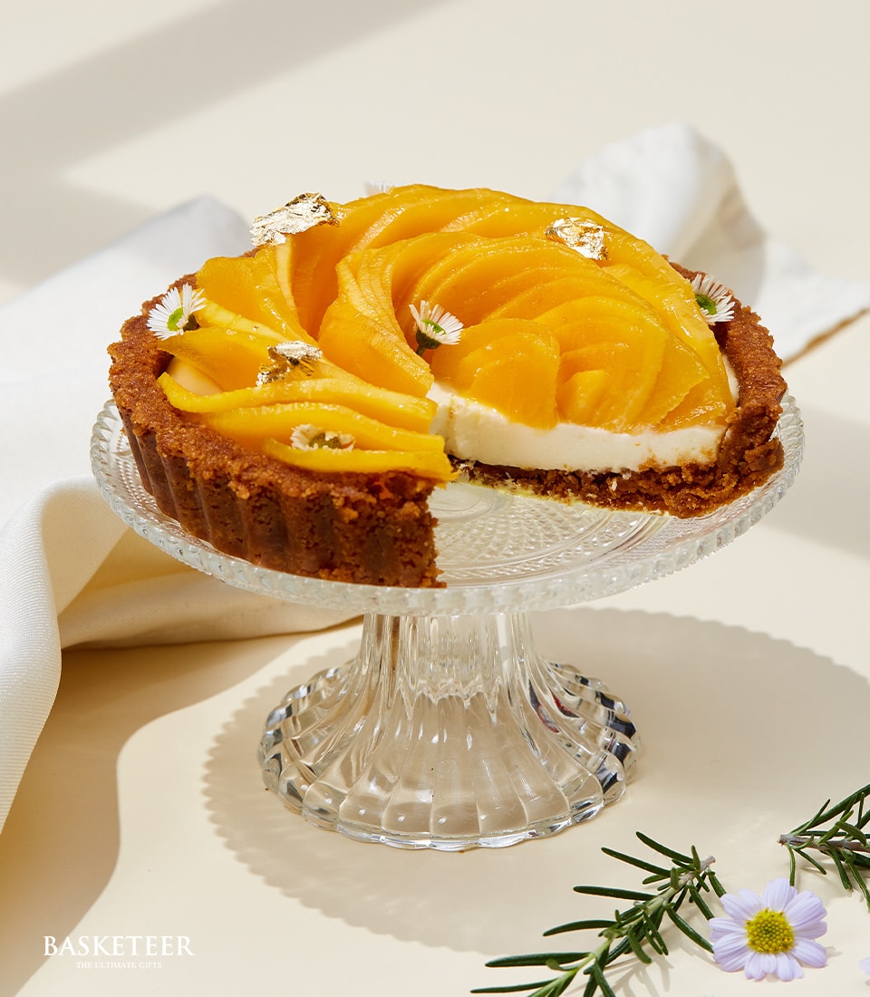 Golden mango cheese tart with candied mango slices and edible flowers on glass cake stand