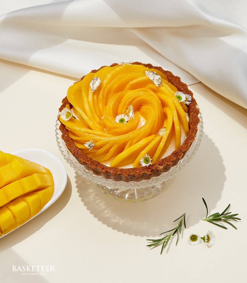 Golden mango tart with spiral fruit topping, white cream filling, and silver leaf garnish on glass stand