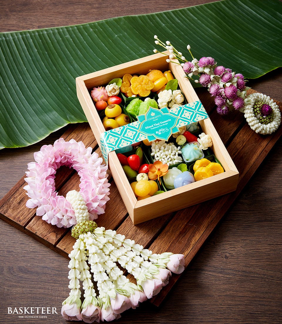 Wooden gift box with colorful Thai desserts and teal ribbon, surrounded by pink and white floral garlands for Songkran