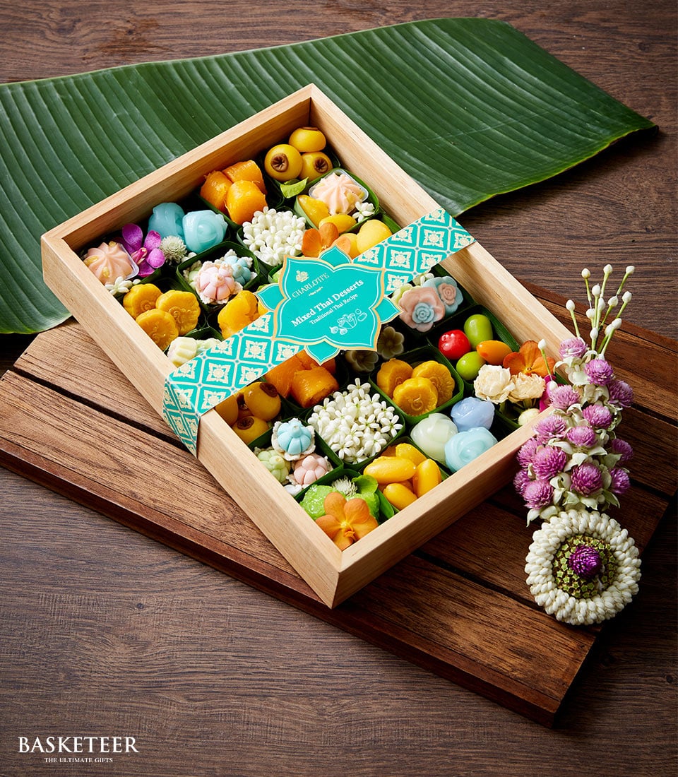 Wooden box of colorful Thai desserts with teal ribbon, banana leaf, and floral garnish for Songkran