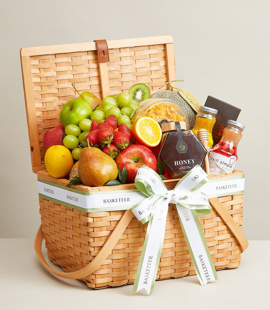 Light brown wicker basket filled with fresh apples, grapes, strawberries, citrus, honey jar, and gourmet treats tied with