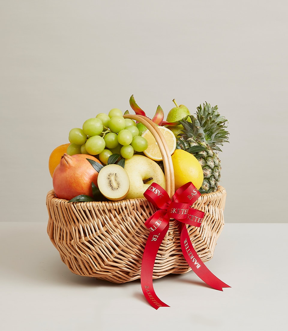 Light brown wicker fruit basket with fresh grapes, kiwi, pomegranate, orange, pear, lemon and pineapple, tied with red ribbon