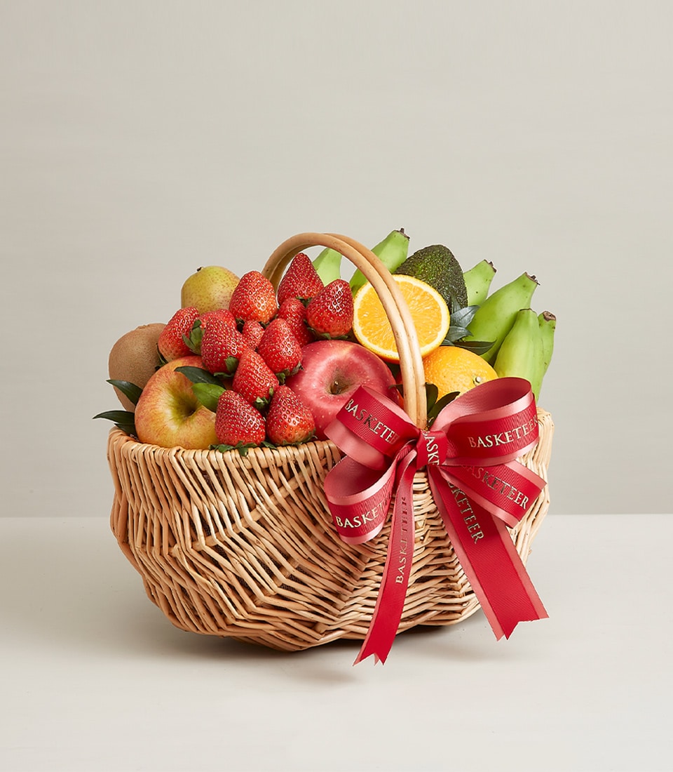 Woven wicker basket overflowing with fresh strawberries, oranges, apples, pears, avocado and bananas, tied with red ribbon