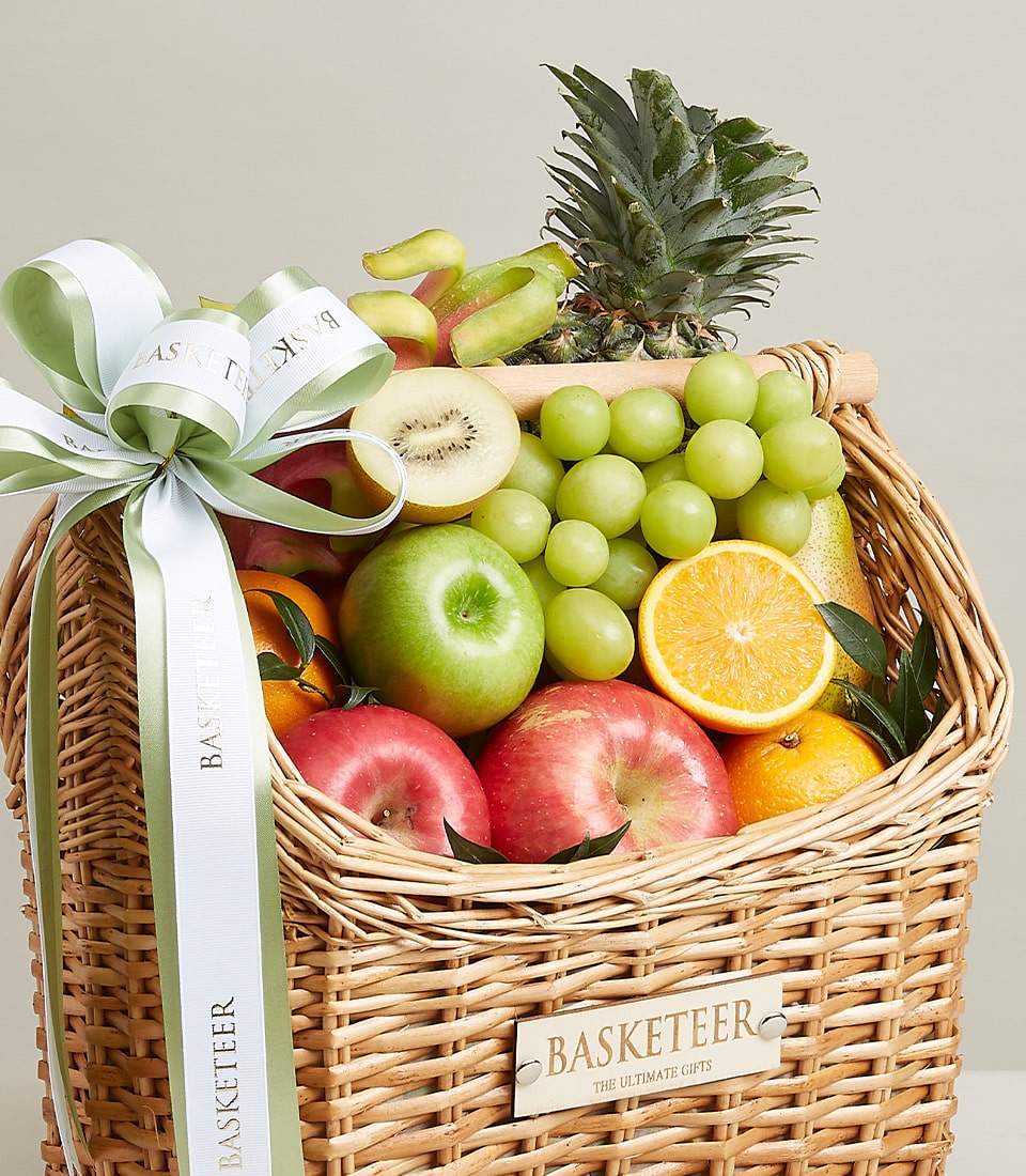 Woven wicker fruit basket filled with fresh apples, grapes, oranges, kiwi and pineapple, tied with green and white ribbon