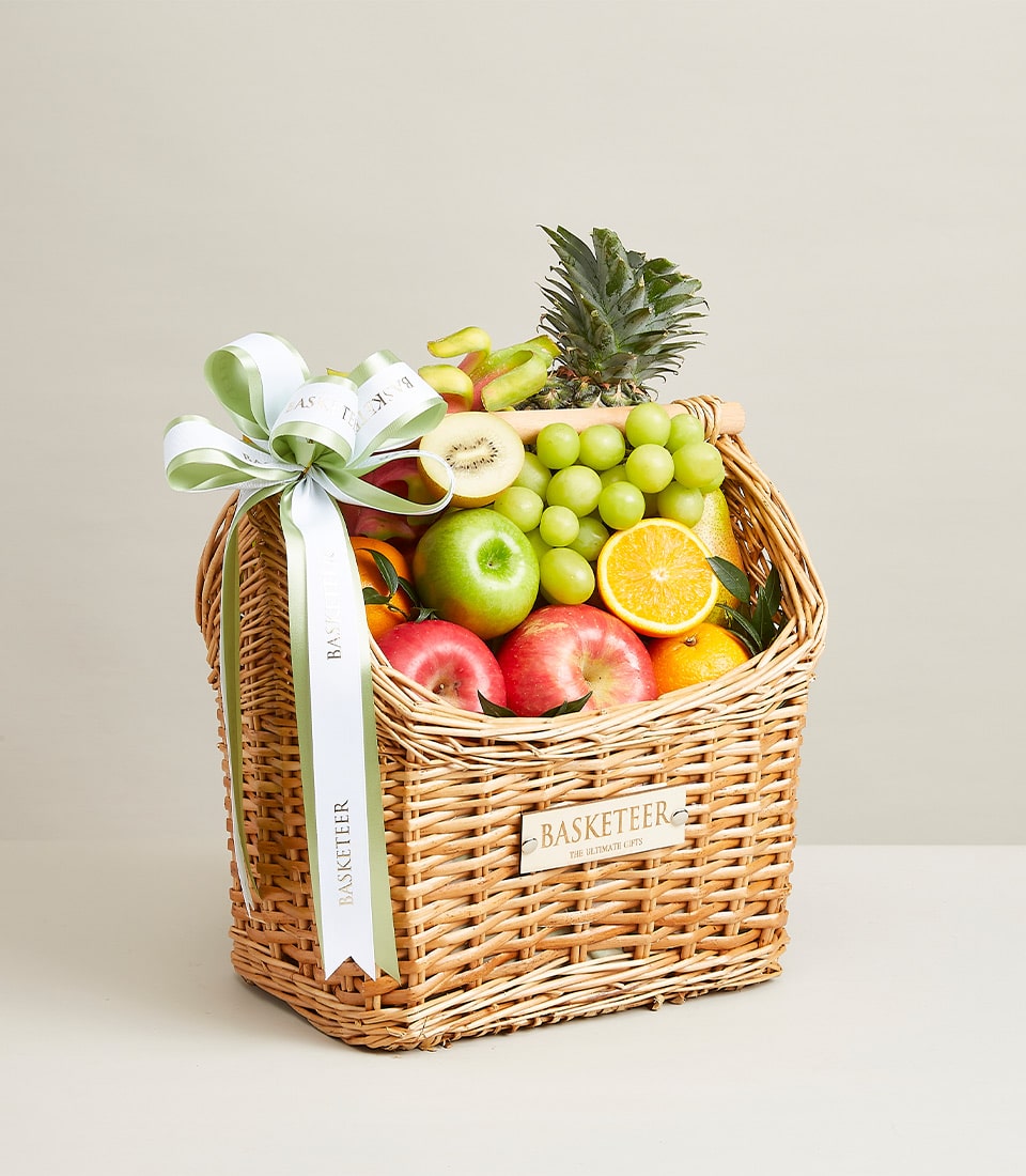 Woven wicker fruit basket with fresh grapes, apples, oranges, kiwi, and pineapple, tied with light green striped ribbon