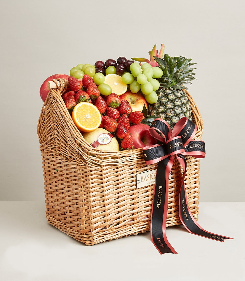 Woven wicker fruit basket with fresh strawberries, grapes, apples, pear, pineapple, dragon fruit and citrus, tied with black