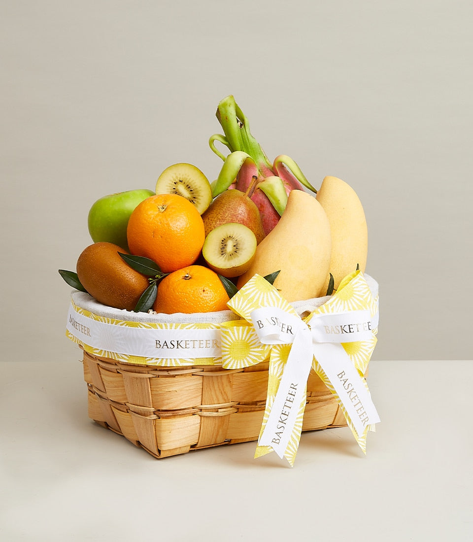 Woven wood gourmet fruit basket with fresh apples, kiwis, oranges, pears, mangoes and dragon fruit, white ribbon bow
