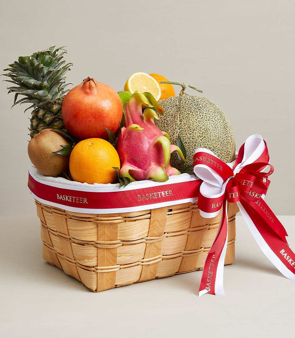 Woven light wood fruit basket with pineapple, pomegranate, dragon fruit, oranges, kiwis and cantaloupe, red and white ribbon