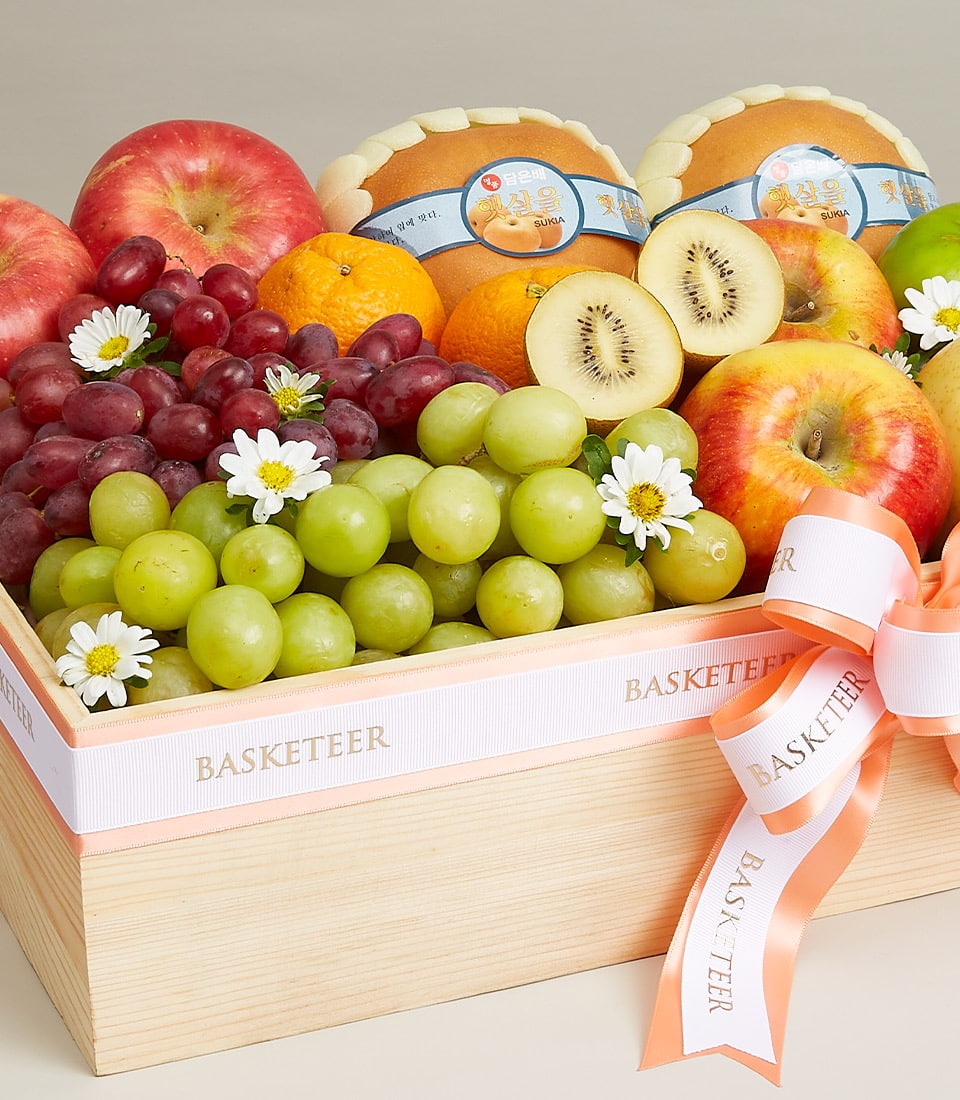 Wooden fruit basket with red apples, green and red grapes, oranges, kiwis, and white flowers with white and peach ribbons