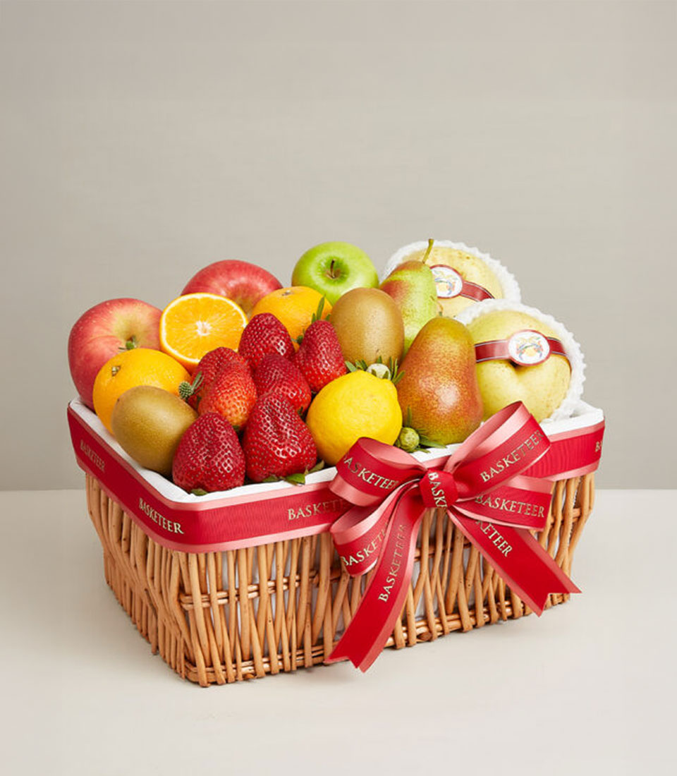 Woven wicker basket filled with fresh mixed fruits, red apples, oranges, kiwis, strawberries and pears, tied with red
