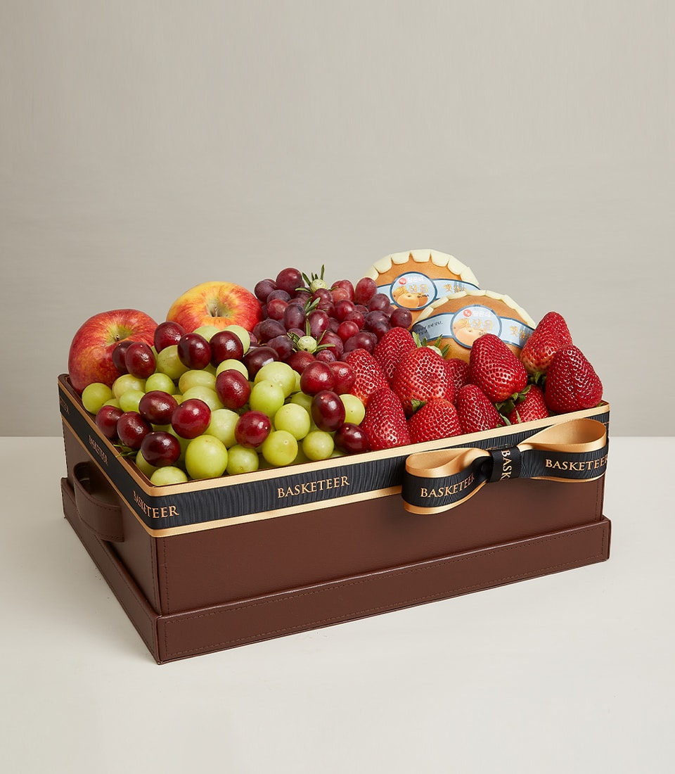 Rectangular brown leather gift box with fresh red apples, grapes, strawberries, and seasonal fruits, tied with black and