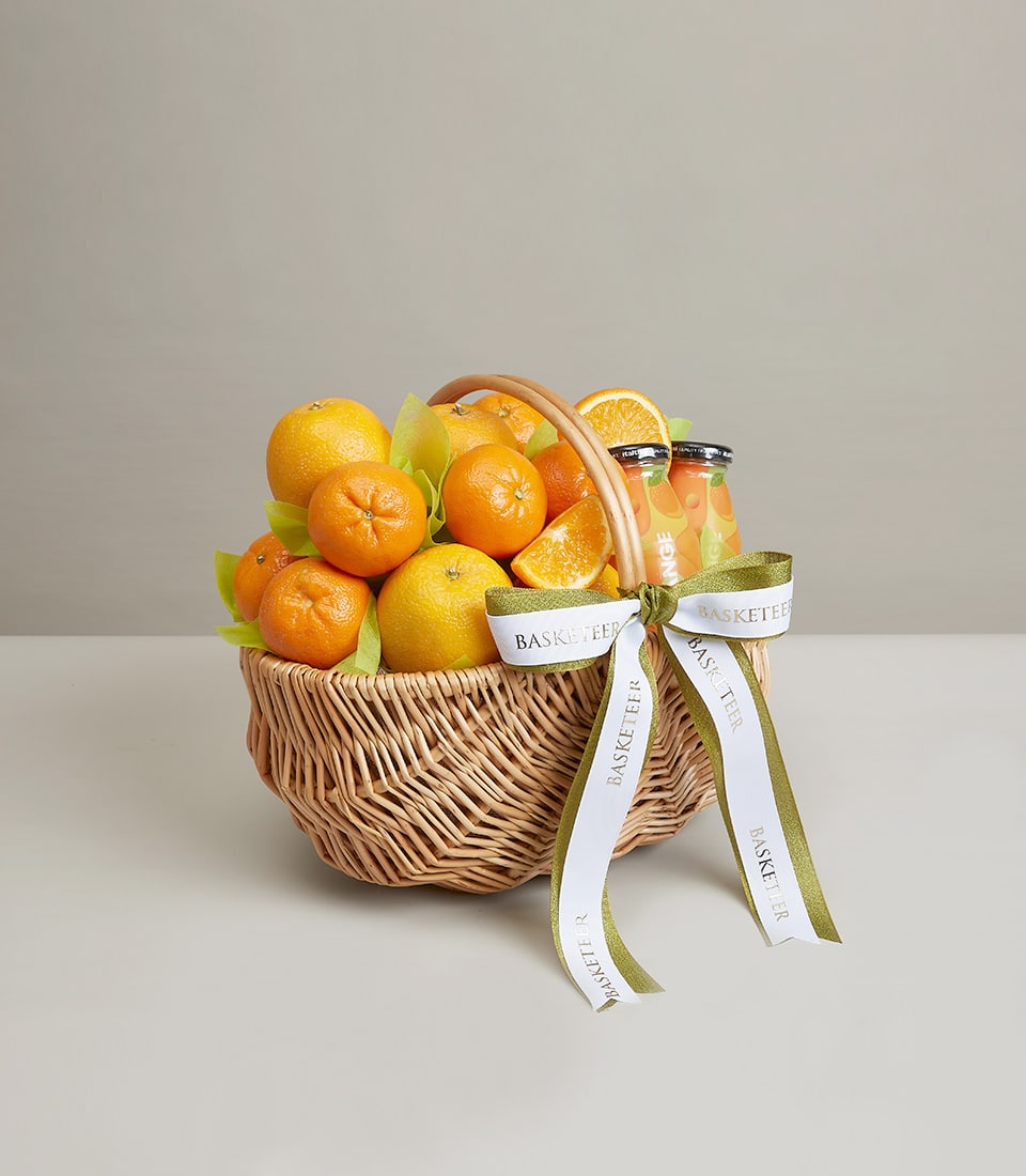 Woven wicker fruit basket with fresh oranges, tangerines, and orange juice bottles tied with white and olive green ribbons